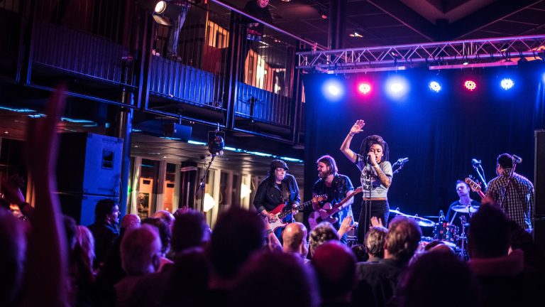 Een band voert op een podium op voor een enthousiaste menigte, verlicht door kleurrijke lichten.