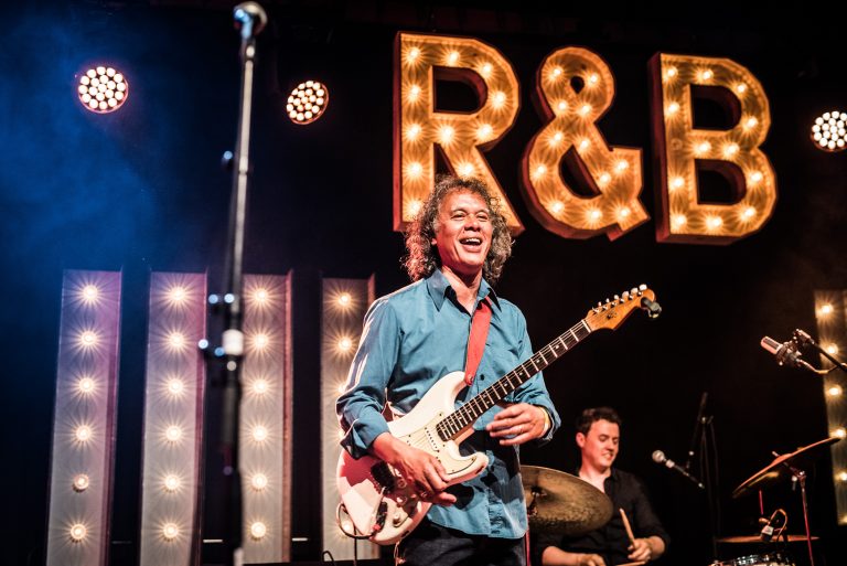 Een man lachend met zijn gitaar in zijn handen met op de achtergrond de letter R&B verlicht