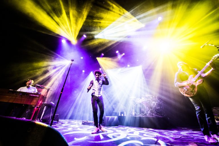 Een muzikant staat op het podium omringd door kleurrijke lichtstralen tijdens een live optreden.