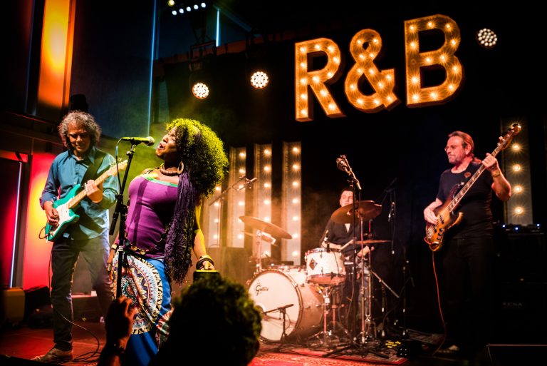 Een muzikale optreden met een zangeres en bandleden op het podium, met een verlichte achtergrond met de tekst