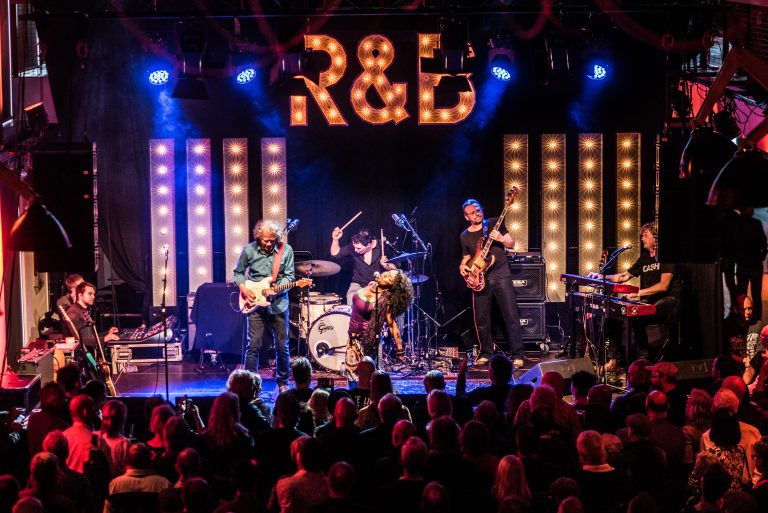 Een band speelt live op het podium, verlicht door blauwe en gouden lichten, met een menigte van fans die naar de voorstelling kijkt.