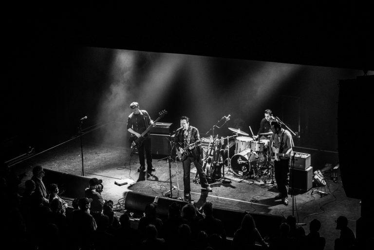 Zwarte-wit afbeelding van een band die live optreedt met muzikanten op een podium en verlichting in de achtergrond.