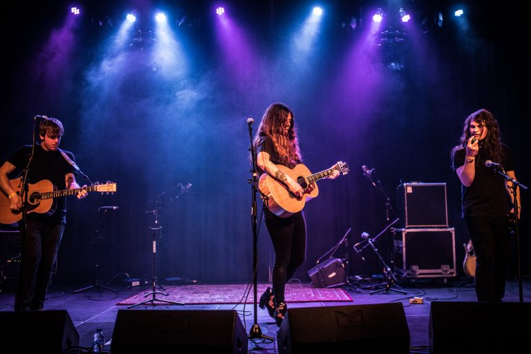 Een muzikante staat op een podium met een gitaar, omringd door kleurrijk licht.