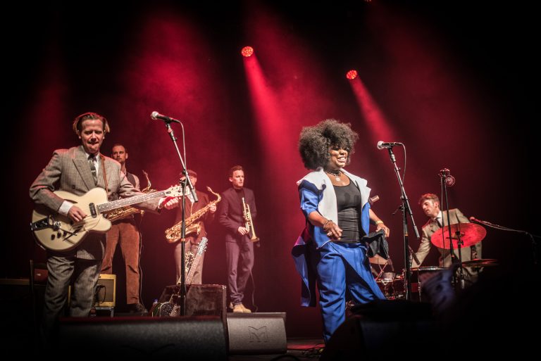 Een energieke liveband op het podium met een vrouwelijke zangeres in een blauw kostuum en muzikanten met instrumenten in een sfeervolle, verlichte omgeving.