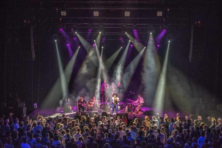 Een live muziekoptreden met bandleden op het podium en een enthousiast publiek onder kleurrijke lichten.