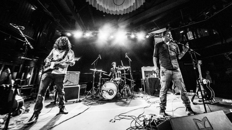 Een band speelt live op het podium, verlicht door felle lichten, met een gitarist, een zanger en een drummer.