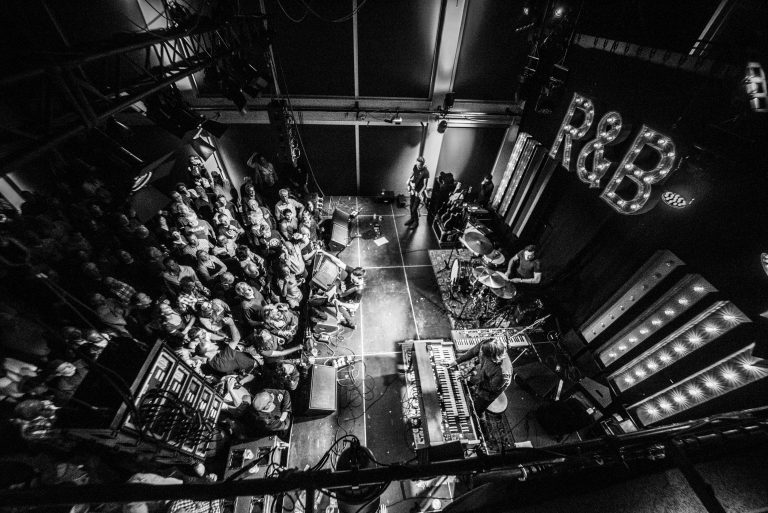 Een bovenaanzicht van een druk concert in zwart-wit, met een publiek dat naar de artiesten op het podium kijkt.