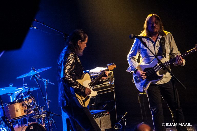 Twee muzikanten voeren een live-optreden uit op een podium, met gitaren en een drumstel op de achtergrond.