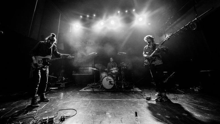 Een zwart-witfoto van een band die live optreedt met een drummer en twee gitaristen op het podium, omringd door lichteffecten.