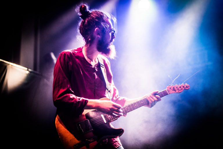Een muzikant speelt gitaar op een podium, omringd door kleurrijke lichtstralen en nevel.