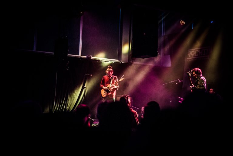 Een muzikant speelt gitaar op een podium, omringd door kleurrijk licht en een publiek.