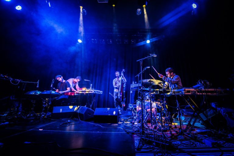 Een live band bestaande uit vier muzikanten die optreden op een podium met verschillende instrumenten en kleurrijke verlichting.