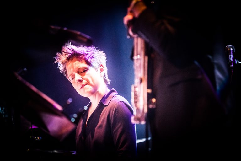 Een muzikant speelt piano op het podium, met een blije uitdrukking en een andere muzikant op de achtergrond.