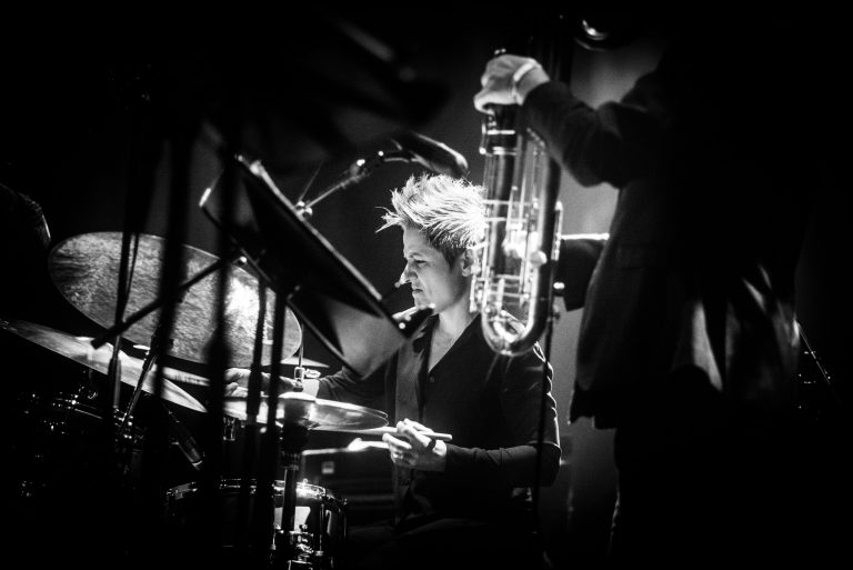 Een drummer in een zwart-wit setting, geconcentreerd aan het werk, terwijl een muzikant met een blaasinstrument op de achtergrond staat.