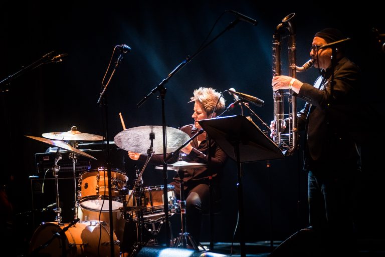 Een drummer en een saxofonist spelen samen op een podium met sfeervolle verlichting.