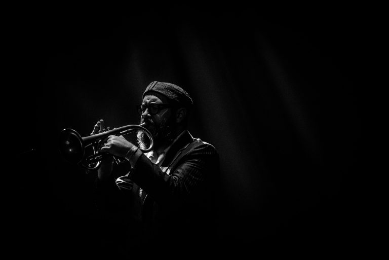 Een muzikant speelt trompet op het podium, verlicht door een enkel lichtpunt in de achtergrond.