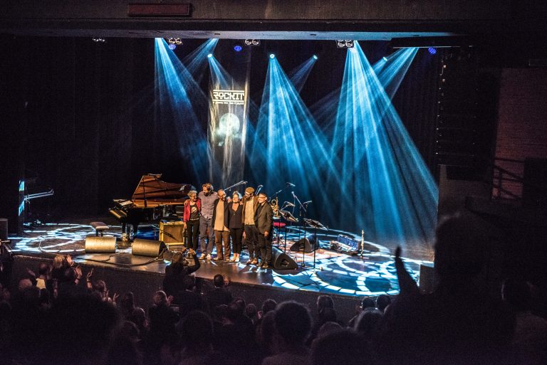 Een groep muzikanten staat op het podium met kleurrijke lichteffecten en een piano op de achtergrond.