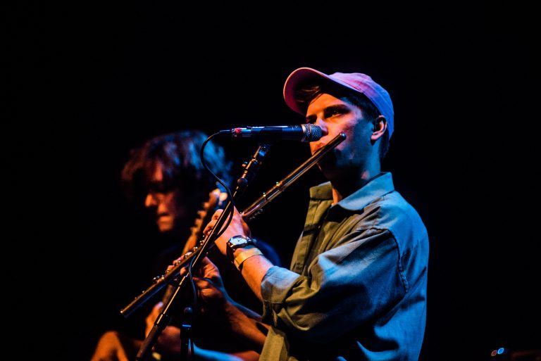 Een muzikant speelt fluit en zingt op het podium, terwijl een andere muzikant op de achtergrond speelt.