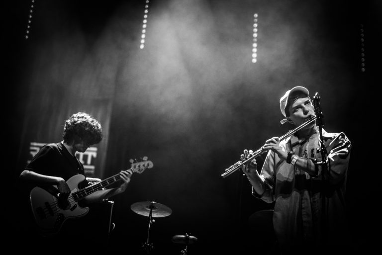 Twee muzikanten performen op een podium, één met een gitaar en de ander met een fluit, omgeven door dim licht en rook.