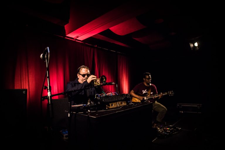 Twee muzikanten voeren een liveoptreden uit met een trompet en een gitaar, verlicht door rode achtergrondverlichting.