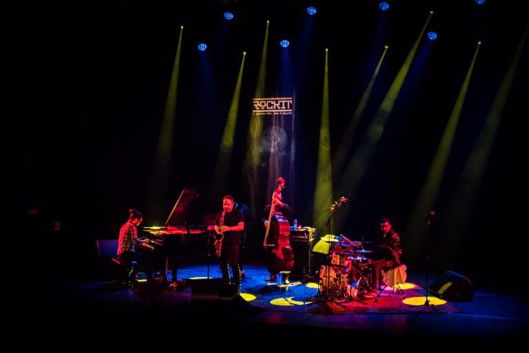 Een band speelt op een podium met kleurrijke verlichting, bestaande uit een pianist, saxofonist, bassist en drummer.