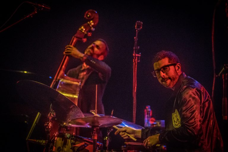 Twee muzikanten spelen samen op een podium, met een contrabas en drums, omgeven door sfeerverlichting.