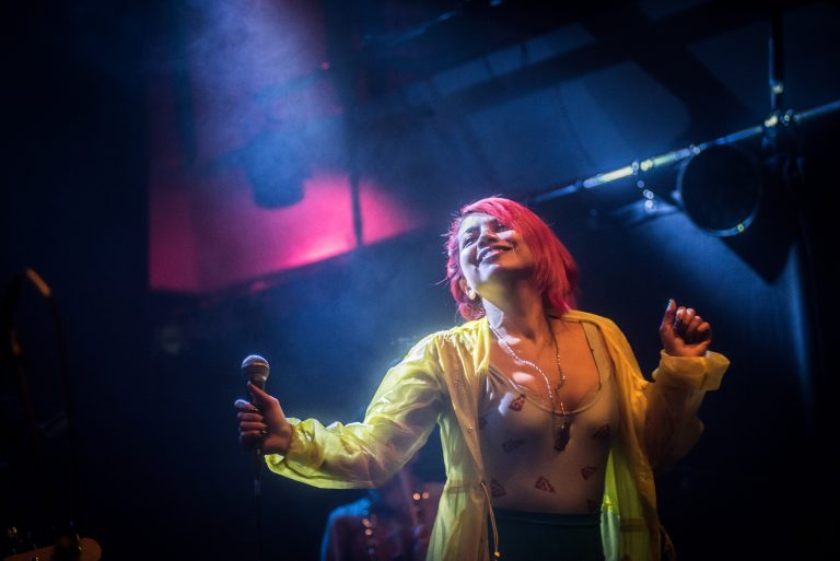 Een energieke performer met een opvallende roze haarkleur zingt op een podium met kleurrijke verlichting.
