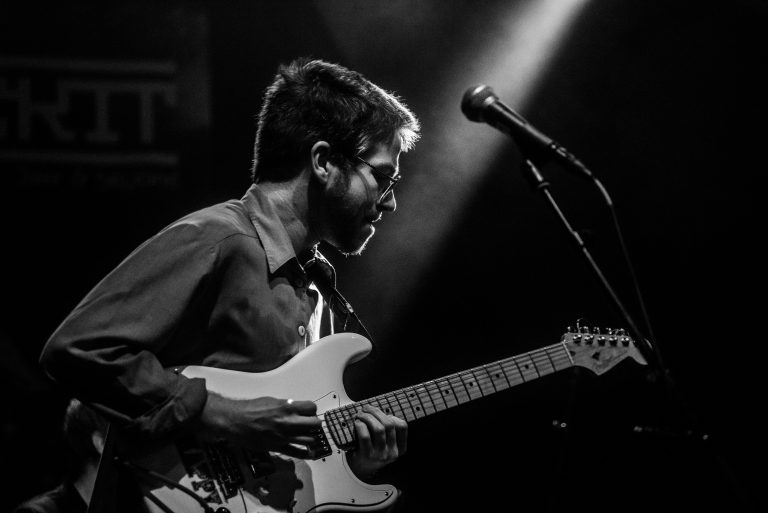 Een muzikant speelt gitaar op een podium, belicht door een enkele lichtstraal.