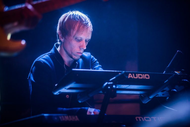 Een muzikant speelt geconcentreerd op een keyboard tijdens een optreden, omgeven door een blauwe lichten.