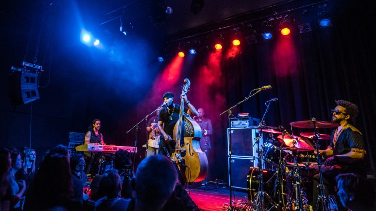 Een bandperformantie op het podium met felgekleurde lichten en verschillende muzikanten die instrumenten bespelen.