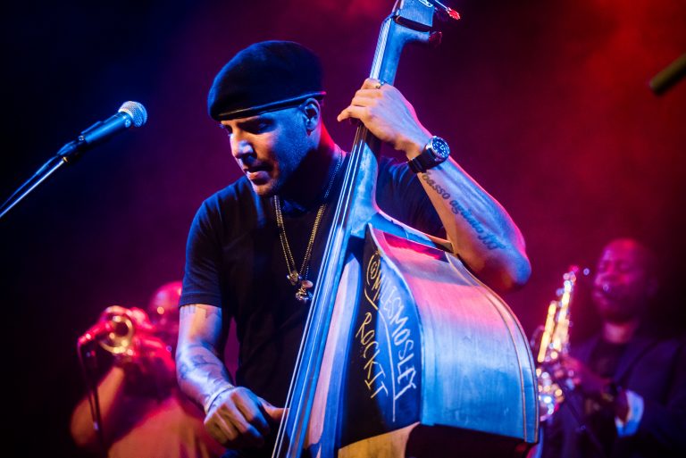 Een muzikant speelt op een contrabas tijdens een optreden, verlicht door kleurige lichten.