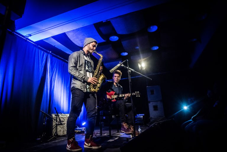 Twee muzikanten optreden op een podium, waarbij één een saxofoon speelt en de ander gitaar speelt, met blauwe verlichting op de achtergrond.