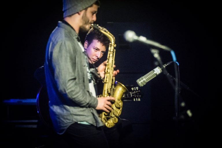 Twee muzikanten op het podium, waarbij de ene op een saxofoon speelt en de andere naar hem kijkt.