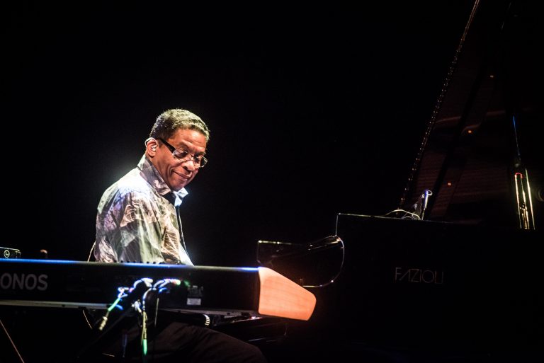 Een man speelt piano op het podium in een donkere setting, met een geconcentreerde blik.