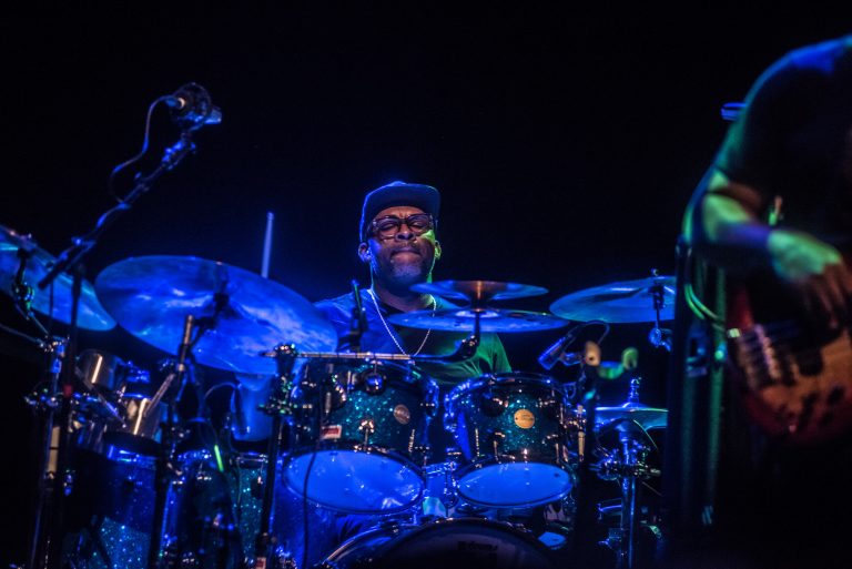 Een drummer in een donker verlichte omgeving, omringd door verschillende trommels en percussie-instrumenten.