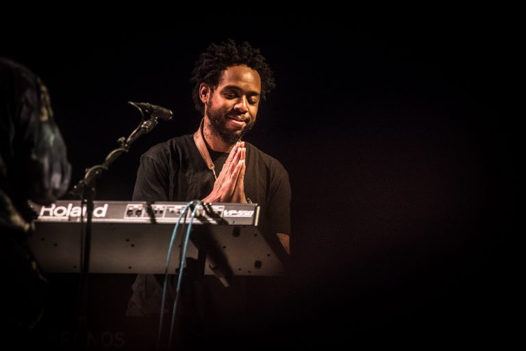 Een muzikant staat achter een keyboard en heeft zijn handen in een gebedshouding.