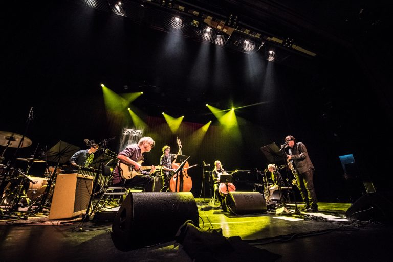 Een liveband bestaat uit zes muzikanten op een podium, omringd door gele en groene lichten.