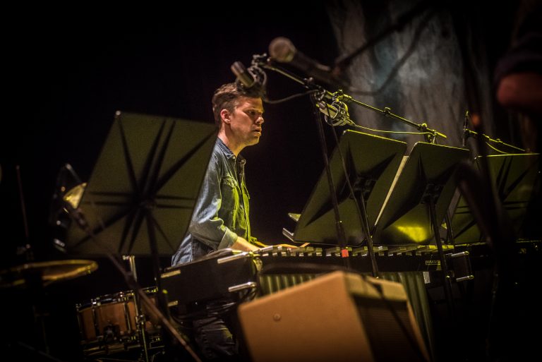 Een muzikant speelt op een keyboard tijdens een live optreden, omringd door muziekstandaards en met een microfoon voor zich.