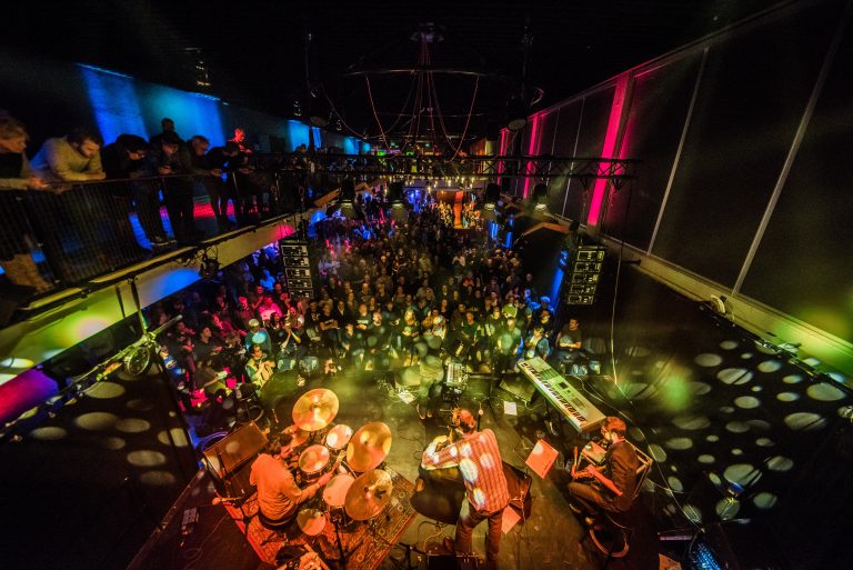 Een druk concert met muzikanten op het podium en een enthousiaste menigte in de zaal, omgeven door kleurrijke verlichting.