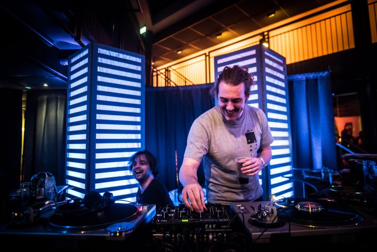 Een DJ staat aan een draaitafel terwijl een andere persoon glimlachend op de achtergrond kijkt, omringd door blauwe en witte verlichting.