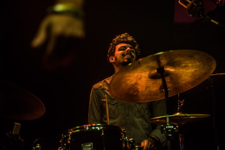 Een drummer speelt enthousiast op een drumstel, met een lichte glimlach op zijn gezicht en theatrale belichting op de achtergrond.