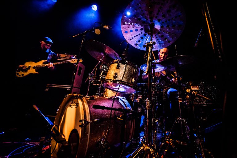 Een drummer speelt op een drumstel met nadruk op de cymbalen, verlicht door blauwe lichten.