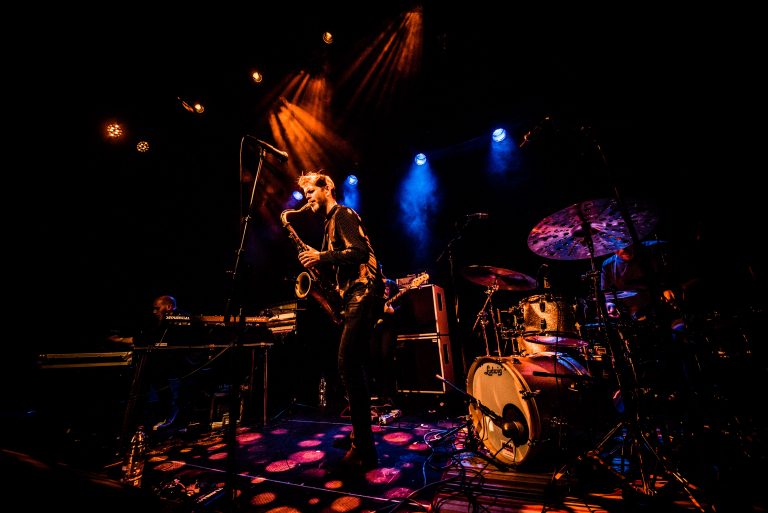 Een muzikant staat op het podium met een saxofon, omringd door instrumenten en kleurrijke lichten.