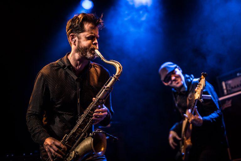 Een muzikant speelt saxofon op een podium, terwijl een andere muzikant op de achtergrond gitaar speelt.
