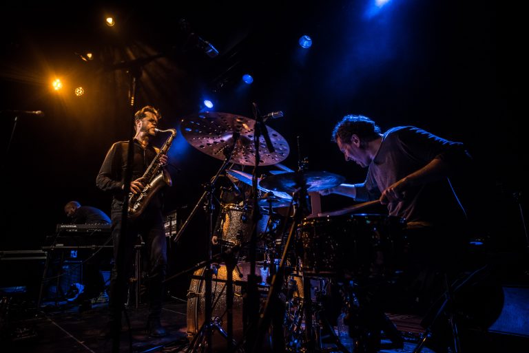 Een saxofonist en een drummer spelen samen op het podium, belicht door blauwe verlichting.