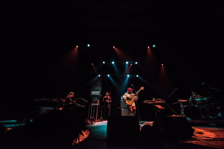 Een band speelt live muziek op een donkere podium met kleurrijke lichten.