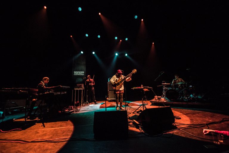 Een muzikant speelt gitaar op een podium, omringd door andere bandleden en sfeervolle verlichting.
