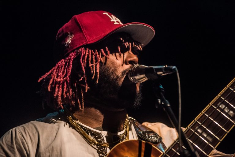 Een muzikant met een rode pet zingt in een microfoon, terwijl hij een gitaar vasthoudt.