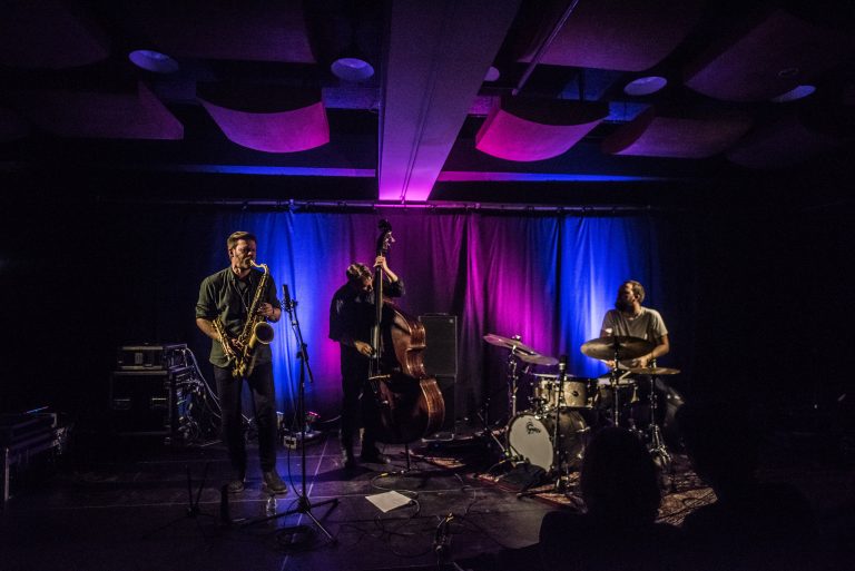 Een jazzband voert een live optreden uit op een sfeervol verlichte podium met paarse en blauwe achtergrond.