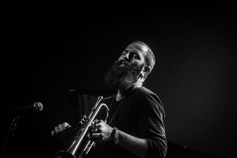 Een muzikant met een baard speelt op een trompet tegen een donkere achtergrond.